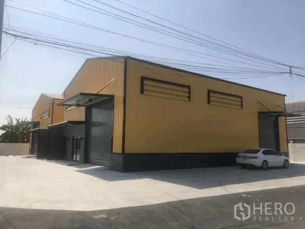 工厂 出租在Bang Yai, Nonthaburi - 相邻厂房侧视，百叶通风口、卷闸门与宽敞混凝土场地。