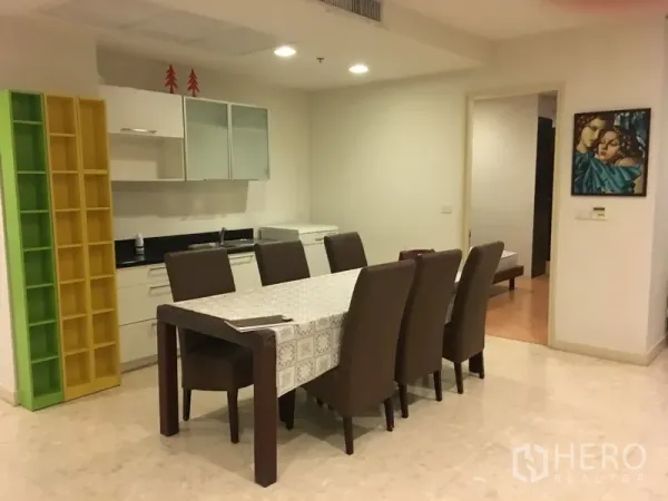 Condominium for rent in Khlong Toei, Bangkok - Dining area with long table for eight beside a compact open kitchenette and white cabinets.