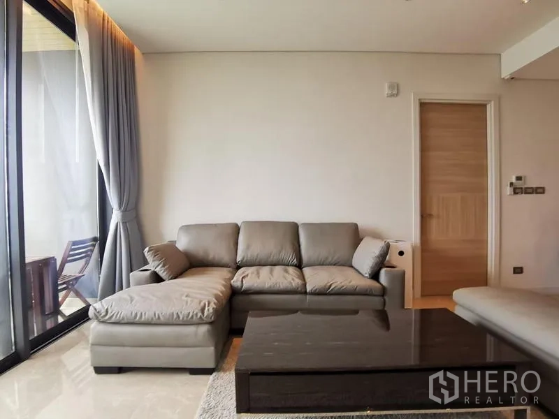 Condominium for rent in Pathum Wan, Bangkok - Cozy lounge with L-shaped sofa beside the balcony and full-height curtains.