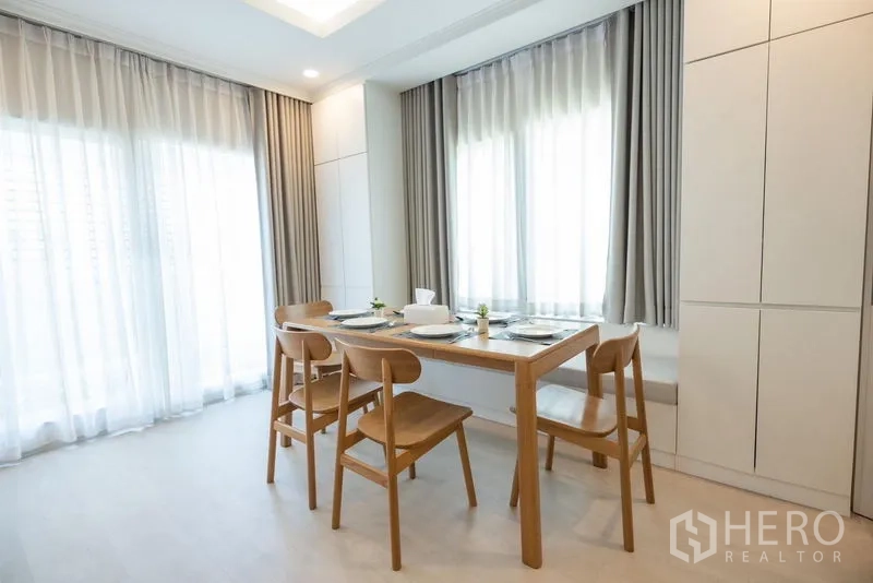 Detached House for sale in Prawet, Bangkok - Dining area with wooden table and four chairs beside full-height windows and sheer curtains.