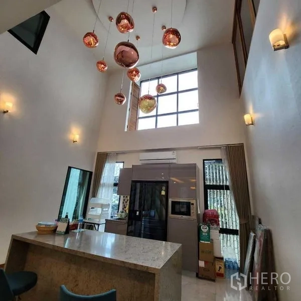 บ้าน ขายในPhra Khanong, Bangkok - ครัวเพดานสูงพร้อมไอส์แลนด์ หน้าต่างสูง และโคมไฟแขวนสีทองแดง