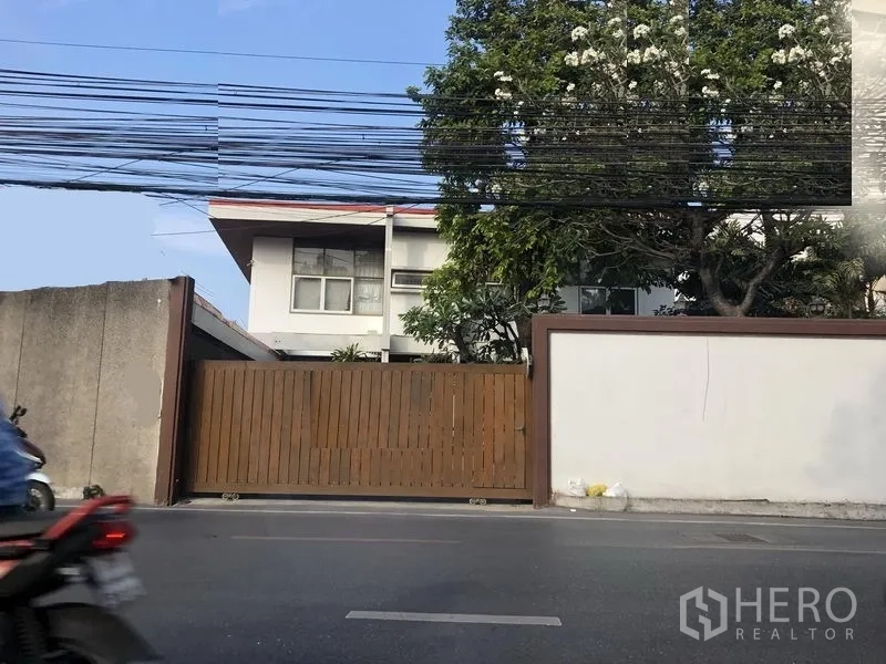 零售空间 出租在Watthana, Bangkok - 街景：艾卡迈区域零售物业正门，白色围墙、木质滑动大门、成熟绿树与两层建筑。