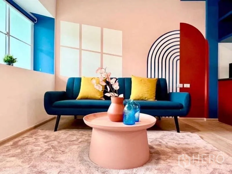 Townhouse for rent in Bang Bo, Samut Prakan - Colorful living room with a teal sofa, yellow cushions, round pastel coffee table and arch mural beside a large window.