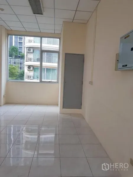 办公空间 出租在Phra Khanong, Bangkok - 明亮转角办公室，落地窗采光，白色地砖，吊顶灯具与配电箱，可见邻近建筑。