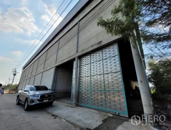Warehouse for rent or sale in Sai Noi, Nonthaburi - Front facade showing a wide sliding metal door and driveway parking beside the warehouse.