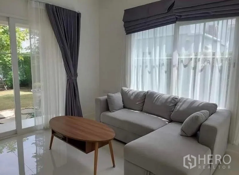 Detached House for rent in Bang Bo, Samut Prakan - Sunlit lounge corner with large windows, gray drapes and a wooden coffee table facing the yard.