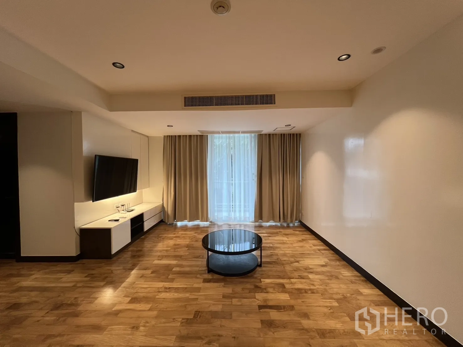 Apartment for rent in Watthana, Bangkok - Spacious living area with wall-mounted TV, built-in console, round coffee table, and full-length curtains.