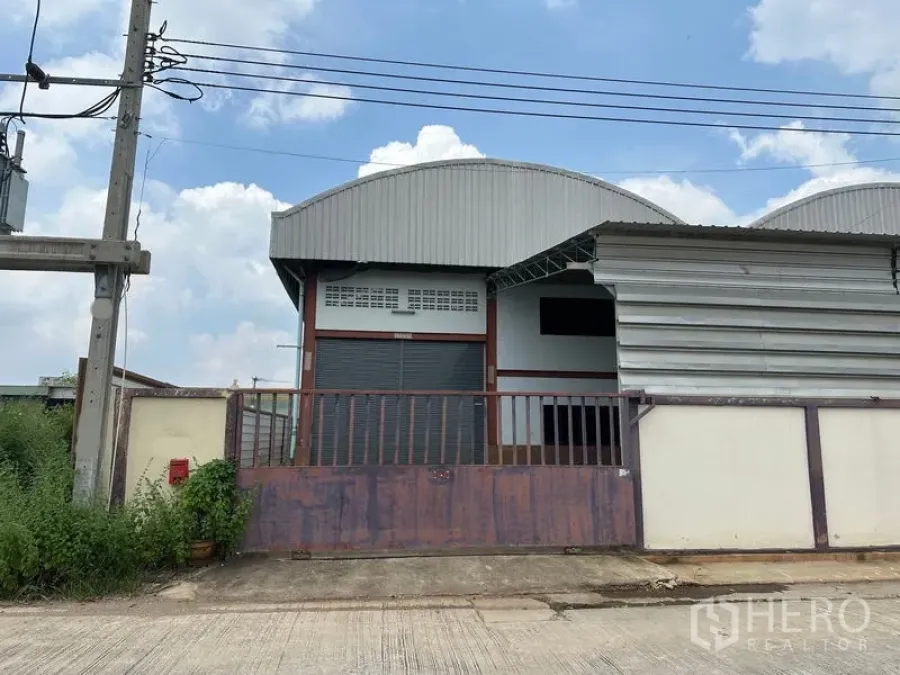 คลังสินค้า ให้เช่าในPak Kret, Nonthaburi - คลังสินค้าติดถนนในปากเกร็ด หลังคาเมทัลชีททรงโค้ง ประตูม้วนกว้าง และประตูรั้วเหล็ก ใต้ท้องฟ้าแจ่มใส