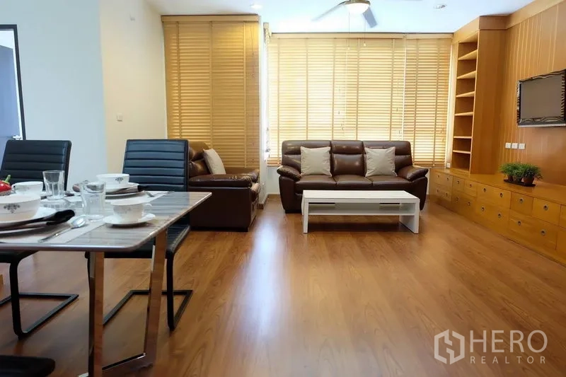 Condominium for rent in Khlong Toei, Bangkok - Open-plan living and dining area with leather sofa set, large windows with blinds, and wood floors