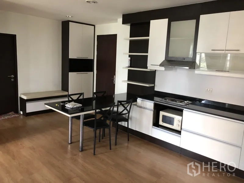 Condominium for rent in Khlong Toei, Bangkok - Open-plan kitchen with white cabinetry, black countertops, cooktop, and dining table on wood floors.