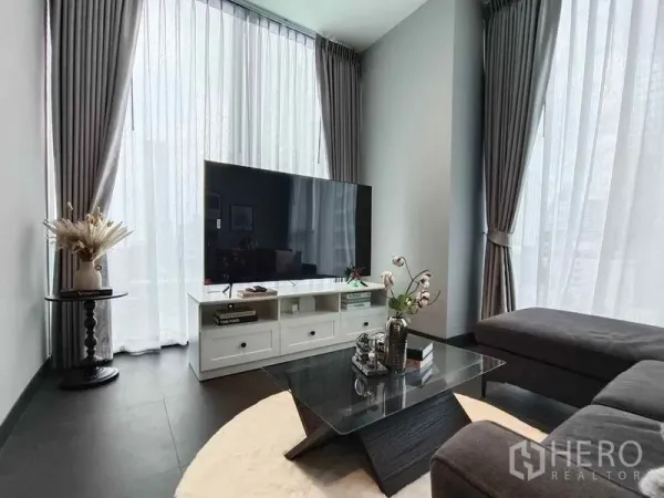 Condominium for rent in Bang Rak, Bangkok - Bright corner living room with sheer curtains, large TV on a white console, and a glass coffee table.