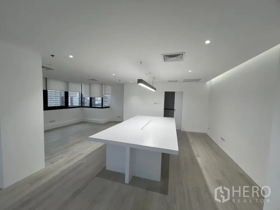 Office Space for rent in Khlong Toei, Bangkok - Bright open‑plan office with long white island table, pendant light, light wood floors and corner windows with roller blinds.