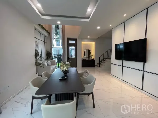 Detached House for rent in Lat Phrao, Bangkok - Open-plan dining zone facing a TV wall and staircase, with recessed ceiling lights and large front windows.