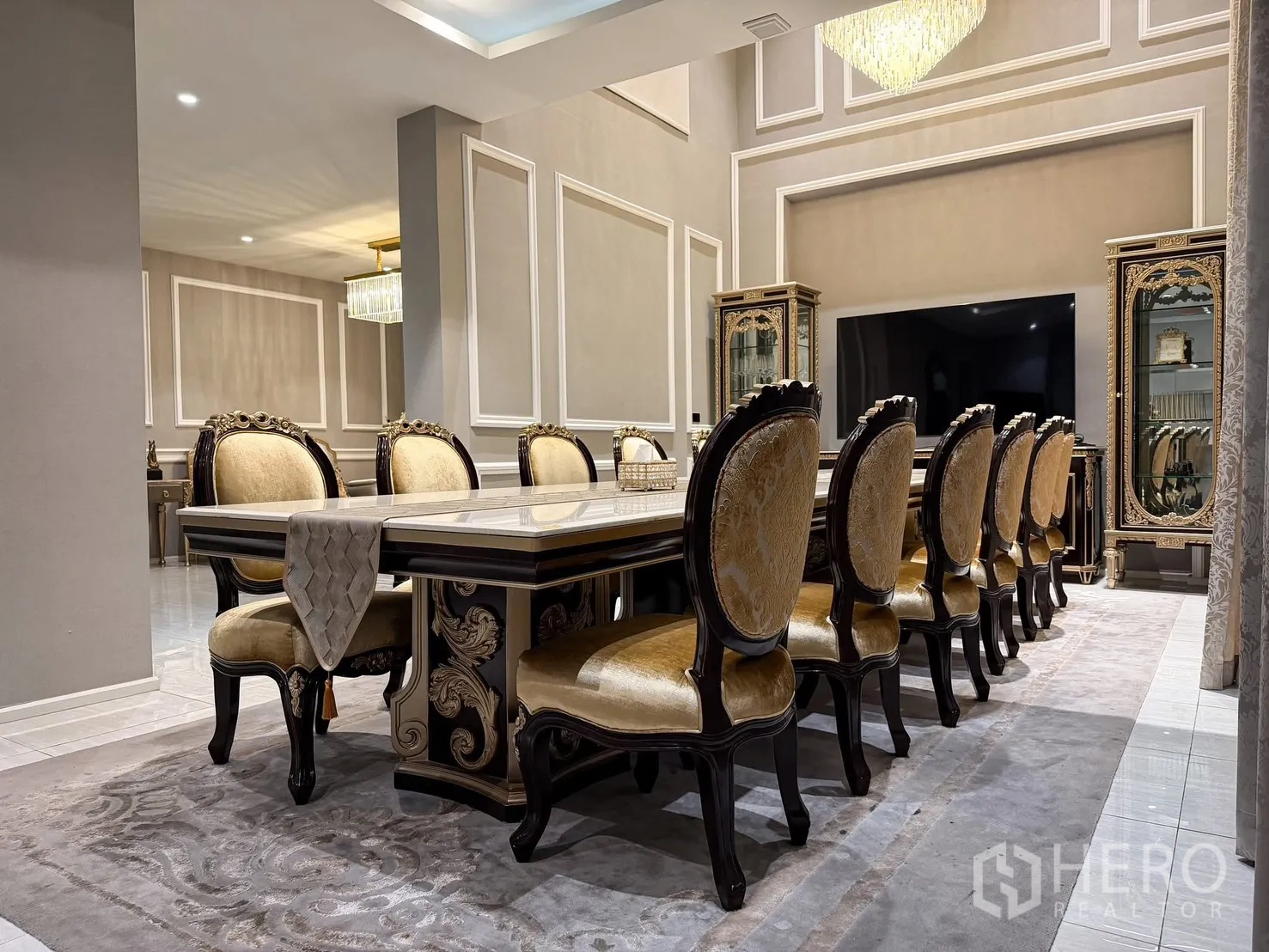 Detached House for rent or sale in Lak Si, Bangkok - Long dining table with many upholstered chairs on an area rug facing a TV wall.