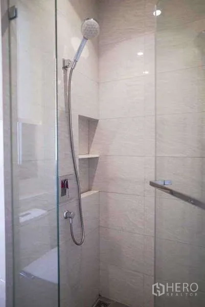 Condominium for rent or sale in Watthana, Bangkok - Glass-enclosed shower with handheld showerhead and built-in wall niches, light stone tiles.