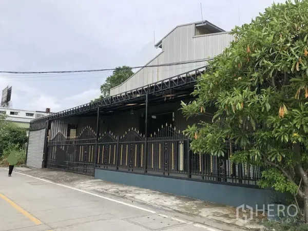 Warehouse for rent in Bang Phli, Samut Prakan - Side view of metal-clad warehouse showing decorative fence and roadside tree.