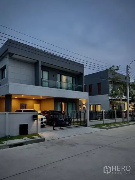 Detached House for rent in Bang Phli, Samut Prakan - Two-storey modern detached house with balcony and gated two-car carport at dusk.