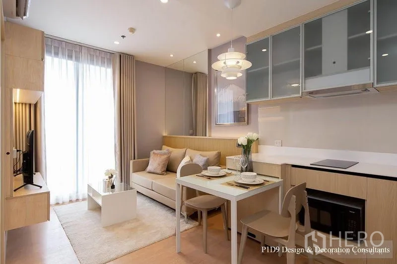 Condominium for rent in Watthana, Bangkok - Living area with balcony doors, mirrored wall, cozy sofa and L‑shaped kitchen beside dining set.