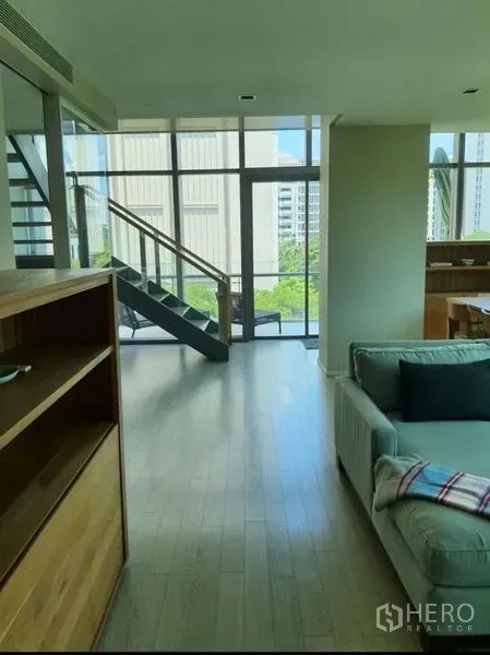 Condominium for rent or sale in Watthana, Bangkok - Loft-style living area with tall windows, light wood floors and a metal-and-glass staircase.