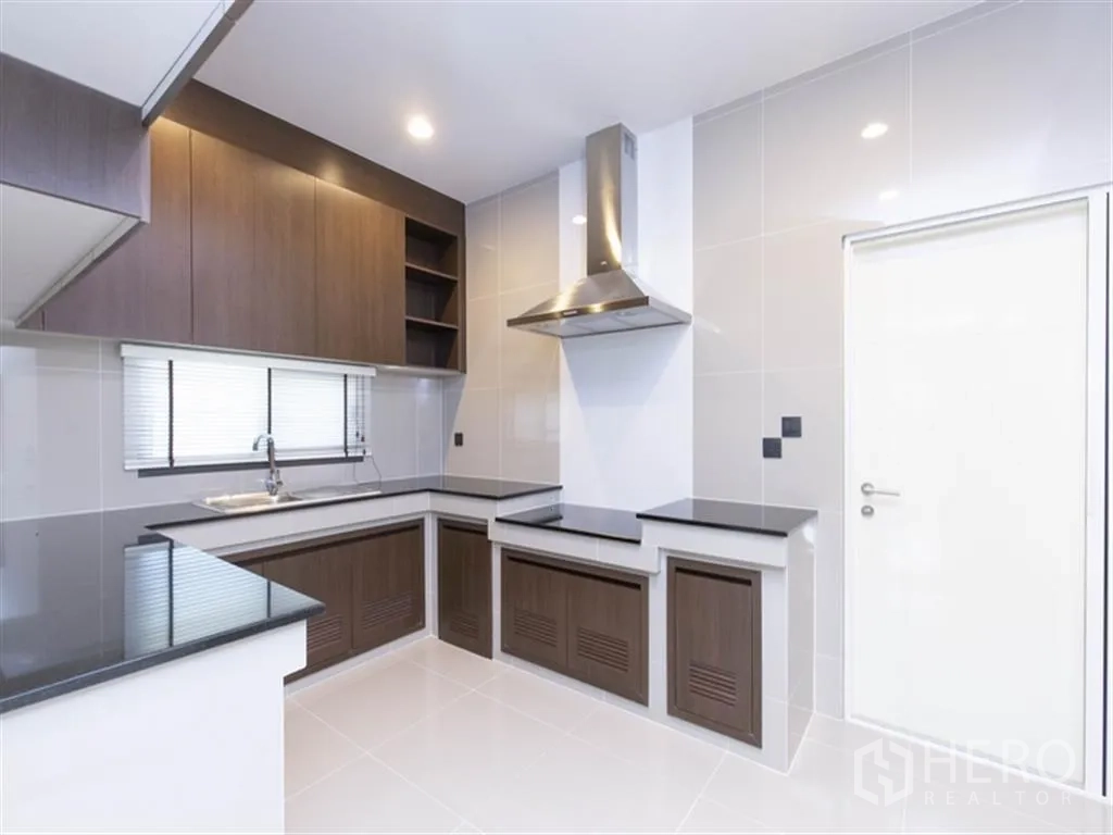 Detached House for rent in Bang Kapi, Bangkok - Modern L-shaped kitchen with dark wood cabinetry, black countertops, window, and stainless range hood.