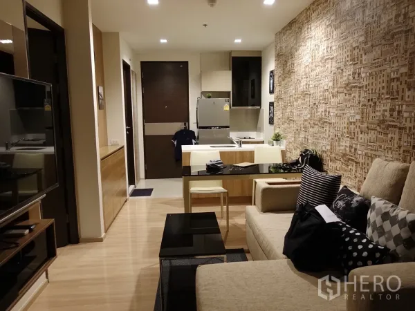 公寓 出租在Sathon, Bangkok - 开放式起居区，可见厨房岛台、电视墙、米色沙发与木地板。