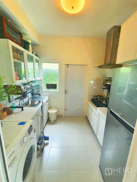 Detached House for sale in Samut Prakan - Compact Thai kitchen with white cabinets, gas hob, range hood and laundry area.
