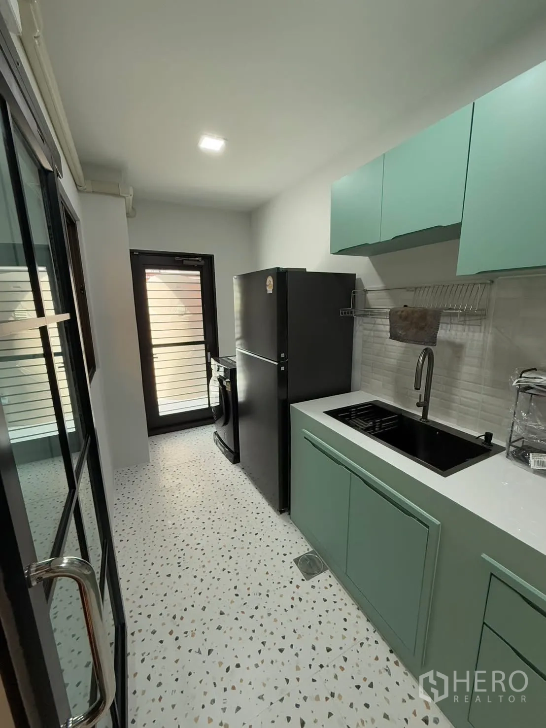 Townhouse for rent in Bang Phli, Samut Prakan - Opposite view of the kitchen showing tall fridge, sink, overhead cabinets and a back door to the utility area.
