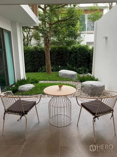 บ้าน ให้เช่าในSaphan Sung, Bangkok - ชานระเบียงมีหลังคา พร้อมเก้าอี้สองตัวและโต๊ะ มองเห็นสวนจัดภูมิทัศน์หินและต้นไม้