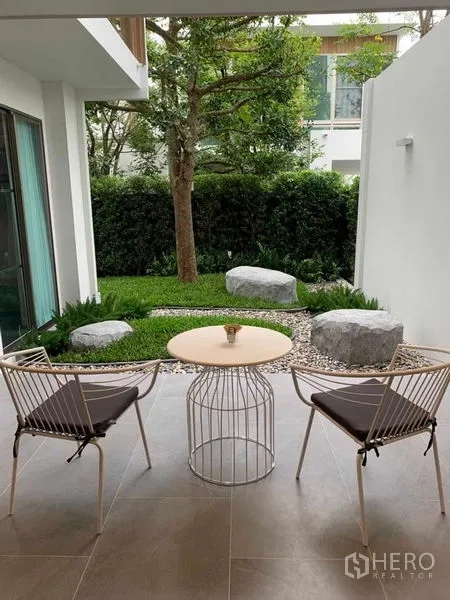 บ้าน ให้เช่าในSaphan Sung, Bangkok - ชานระเบียงมีหลังคา พร้อมเก้าอี้สองตัวและโต๊ะ มองเห็นสวนจัดภูมิทัศน์หินและต้นไม้