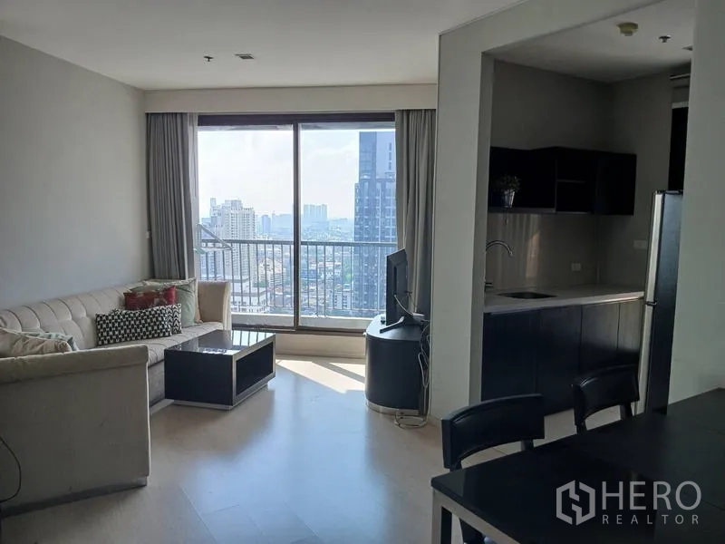 คอนโด ให้เช่า - ห้องนั่งเล่นสว่าง โซฟา ทีวี และระเบียงชมวิวสกายไลน์กรุงเทพฯ ติดครัวแบบเปิด