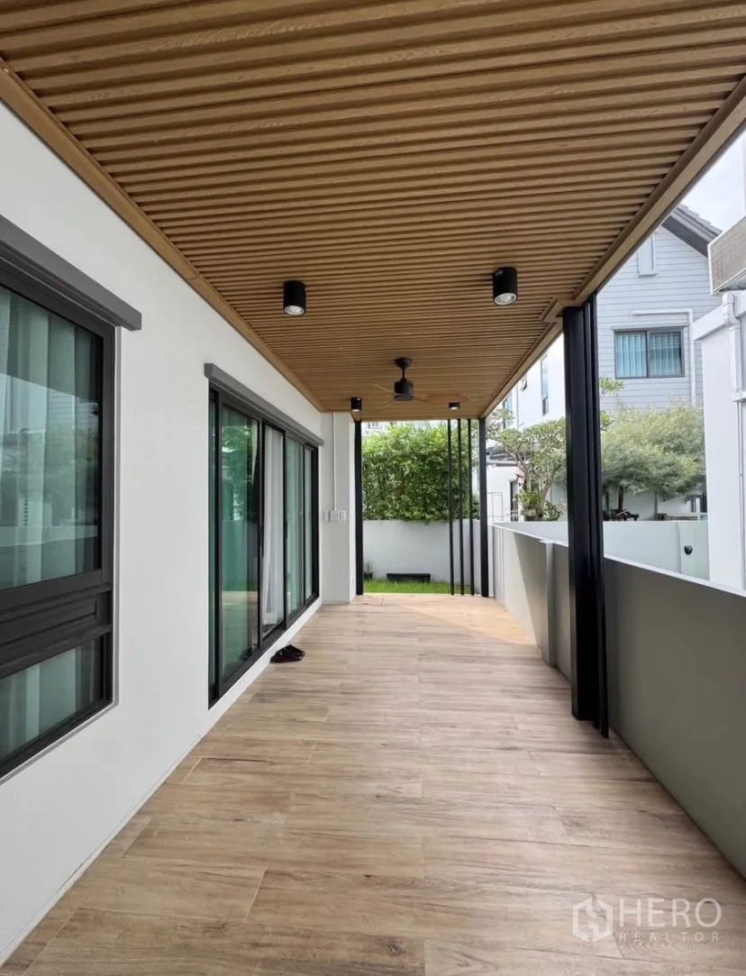 บ้านเดี่ยว for rent or sale in Lat Krabang, Bangkok - ระเบียงทางเดินแบบมีชายคา ปูพื้นลายไม้ เพดานระแนง และประตูกระจกบานเลื่อน เปิดสู่สนามหญ้าเล็กๆ