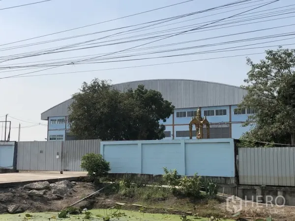 工厂 出售在Lat Lum Kaeo, Pathum Thani - 蓝色厂房外观，拱形屋顶与围墙，前方有运河与树木