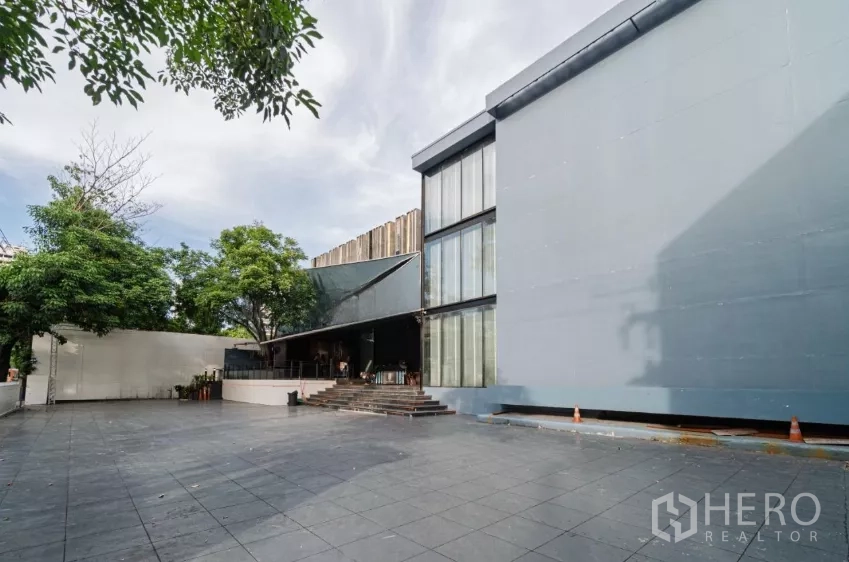 独立商业地产 出租在Watthana, Bangkok - 瓦他那现代独栋建筑，玻璃立面与宽阔铺装前庭并有绿树遮荫。