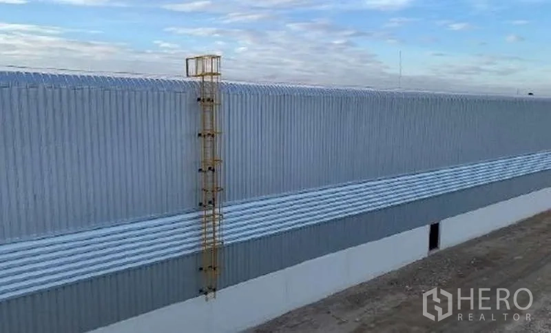 货栈 出租在Mueang Rayong, Rayong - 仓库侧立面，金属波纹墙体外加安全爬梯，天空晴朗。