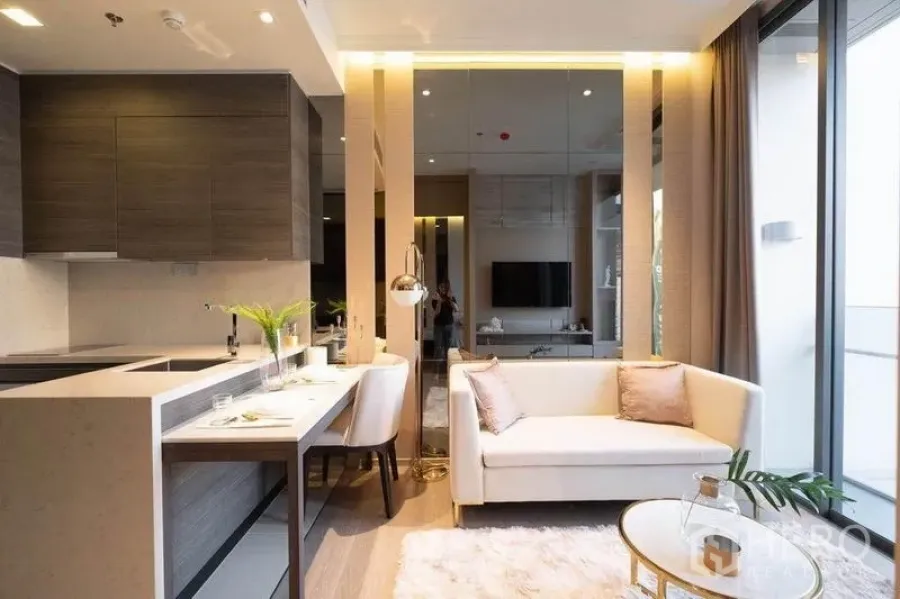 Condominium for rent in Watthana, Bangkok - Bright living area with beige sofa, round coffee table and floor-to-ceiling windows beside a compact kitchen.