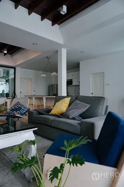 Townhouse for rent in Watthana, Bangkok - Comfortable living room with gray sofa and blue armchair facing an open kitchen beneath exposed beams.