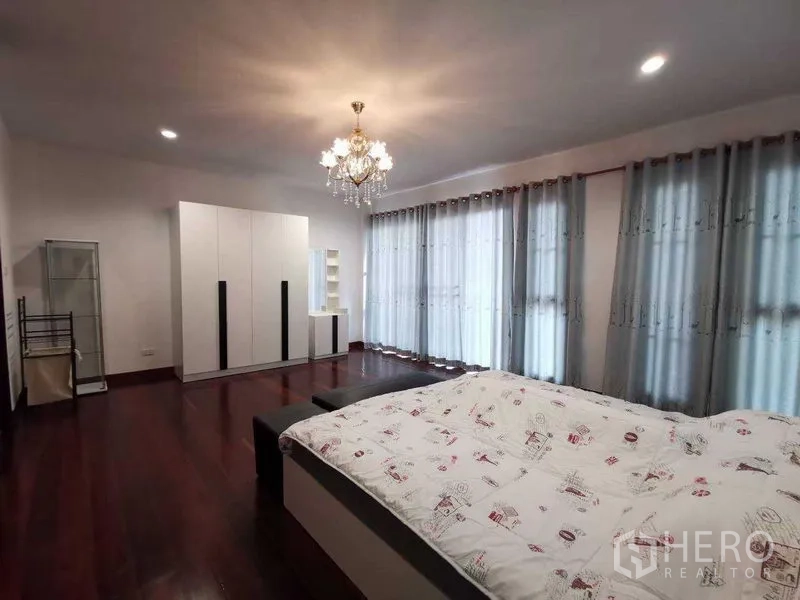 Detached House for rent in Prawet, Bangkok - Spacious bedroom with chandelier, wall of windows, grey curtains and white wardrobes on hardwood flooring.