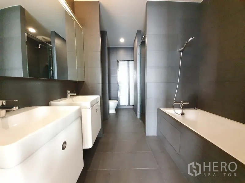 Condominium for rent in Pathum Wan, Bangkok - Luxury bathroom with double vanity, bathtub and a separate glass shower in dark tile finish.