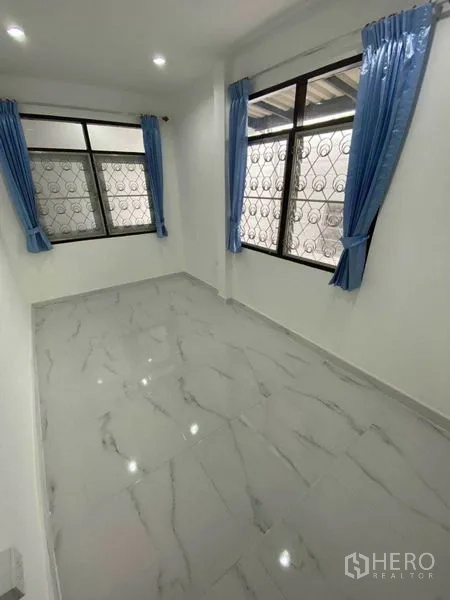 Detached House for rent in Huai Khwang, Bangkok - Sunlit room with blue curtains and security grilles on large windows.