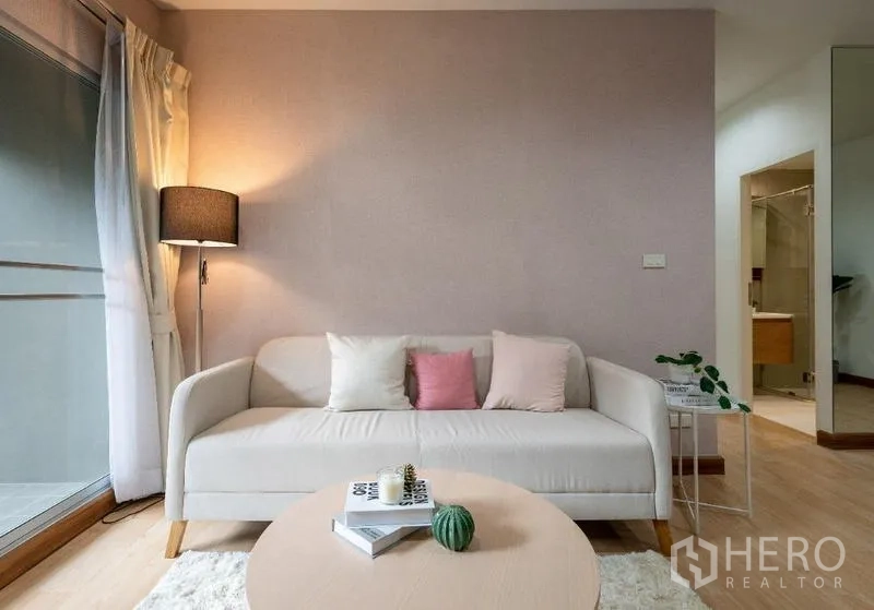 คอนโด ขาย - มุมห้องนั่งเล่น โซฟาโทนอบอุ่น โต๊ะข้าง โคมไฟ และประตูระเบียง