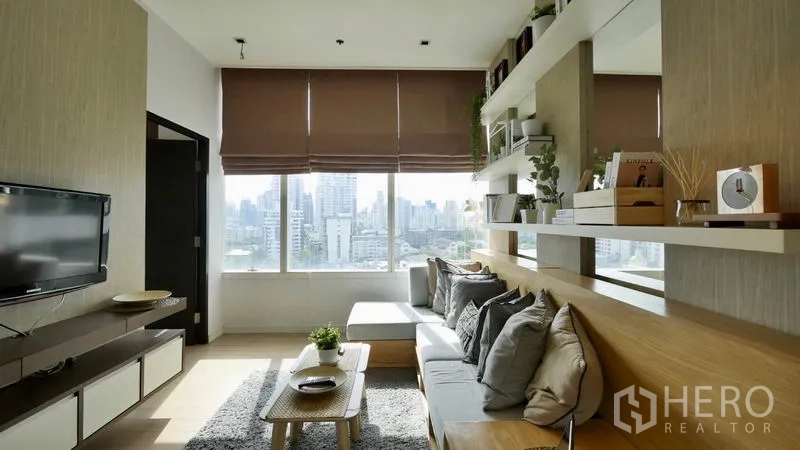 Condominium for rent or sale in Khlong Toei, Bangkok - Living room with large window showcasing Bangkok city skyline, wall-mounted TV and shelving.