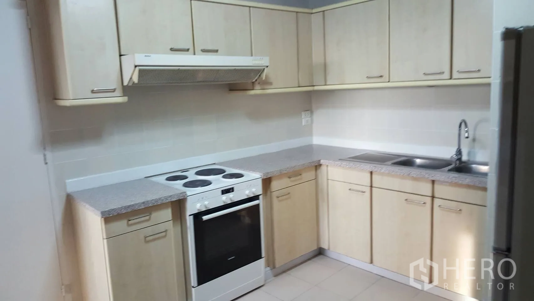 Condominium for rent in Khlong Toei, Bangkok - L-shaped kitchen with light wood cabinets, electric stove and oven, double sink and tiled floor.