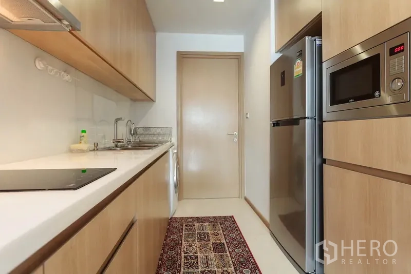 คอนโด ให้เช่าในWatthana, Bangkok - ครัวแบบแกลเลอรี ตู้ลายไม้ เคาน์เตอร์สีขาว เตาอินดักชัน ตู้เย็น ไมโครเวฟ และเครื่องซักผ้า
