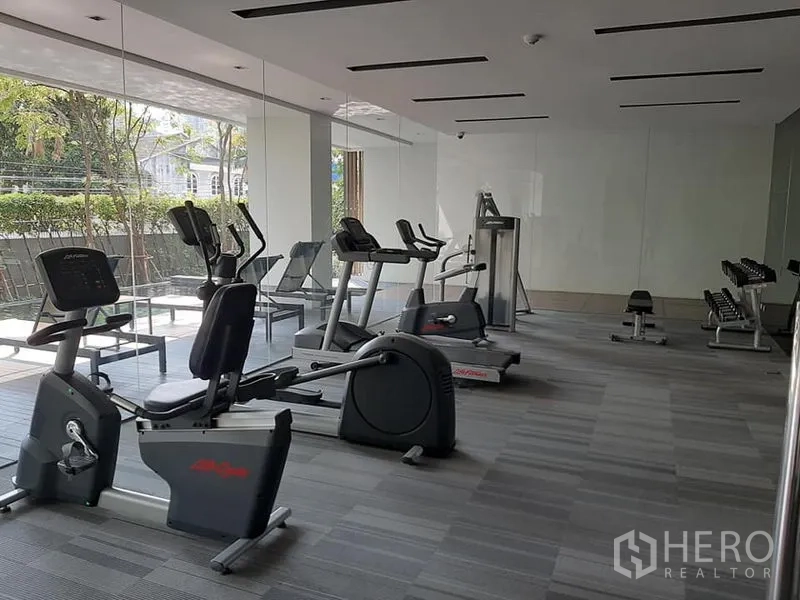 公寓 出售在Watthana, Bangkok - 公寓健身房，跑步机、脚踏车与哑铃，玻璃墙面临绿篱。