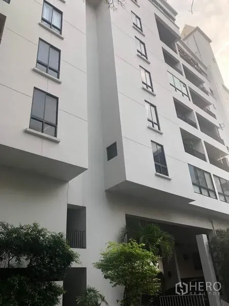 คอนโด ขาย - มุมมองภายนอกคอนโดมิเนียมสีขาวแบบโลว์ไรซ์ มีระเบียงและต้นไม้รอบอาคาร