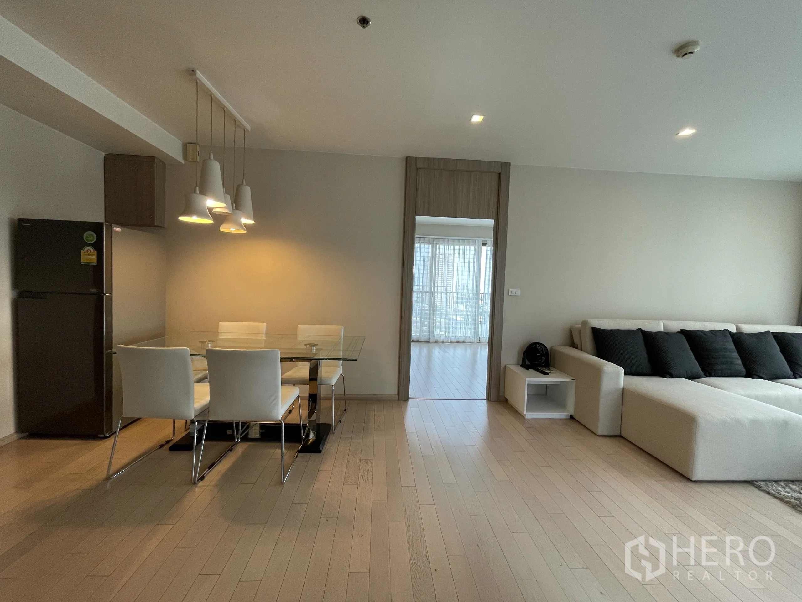 公寓 出租在Watthana, Bangkok - 开放式客餐厅，玻璃餐桌与吊灯，L 型沙发，通往卧室的入口。