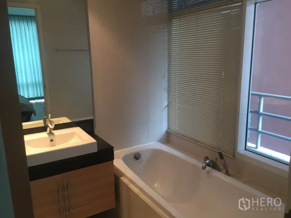 Condominium for rent or sale in Watthana, Bangkok - Bathroom featuring a long soaking bathtub beside a window and a vanity with mirror.