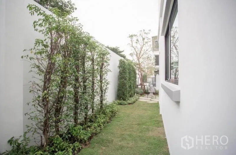 Townhouse for rent in Bang Phli, Samut Prakan - Side garden with lawn, tall hedge and white boundary wall.