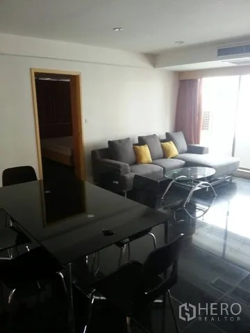 公寓 出租在Bang Rak, Bangkok - 开放式客厅，灰色沙发搭配黄色抱枕，玻璃茶几与餐桌，旁有阳台门。