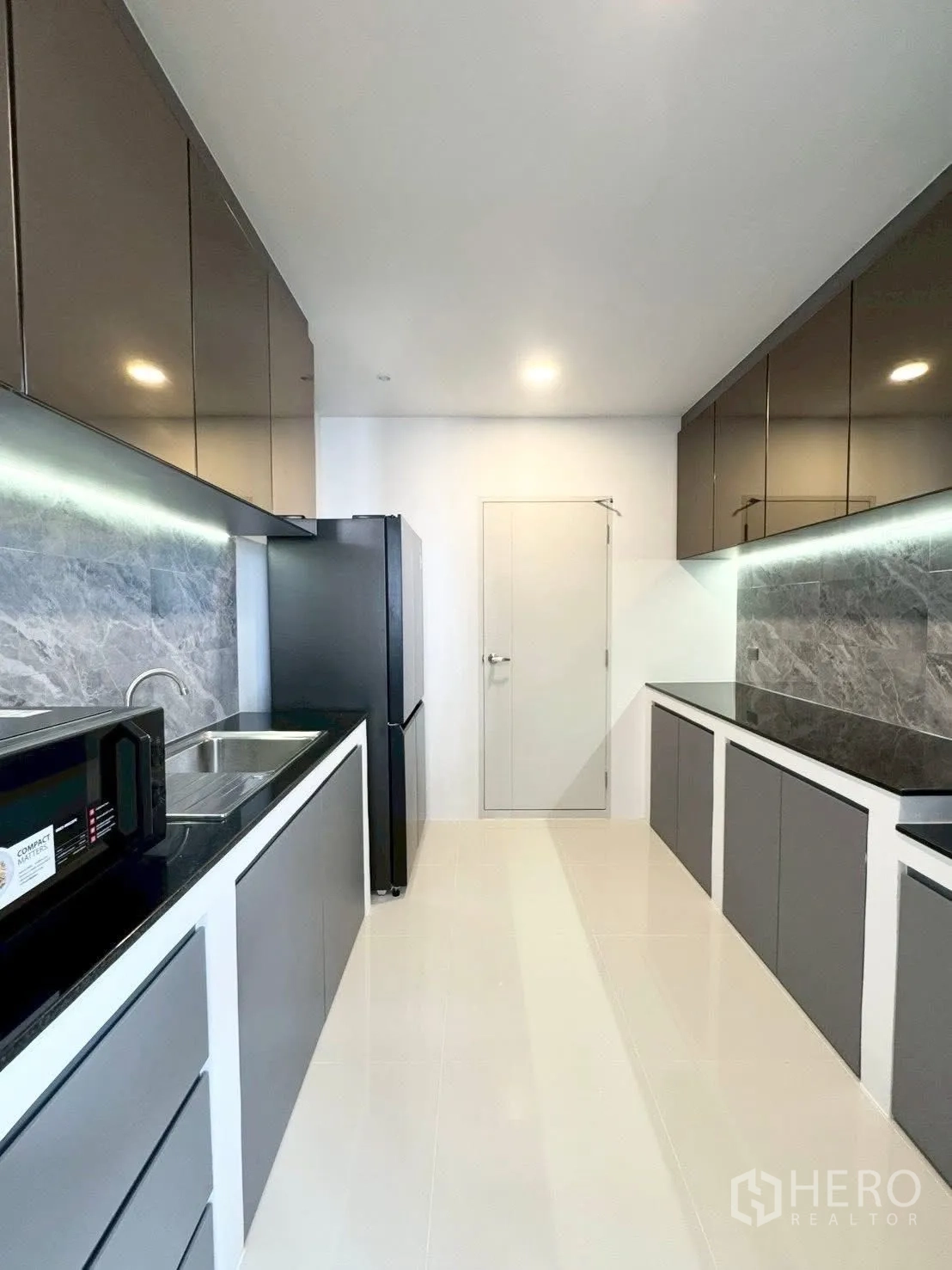 Detached House for rent in Bang Phli, Samut Prakan - Modern galley kitchen with grey cabinetry, black countertops, tile floor, fridge and microwave.